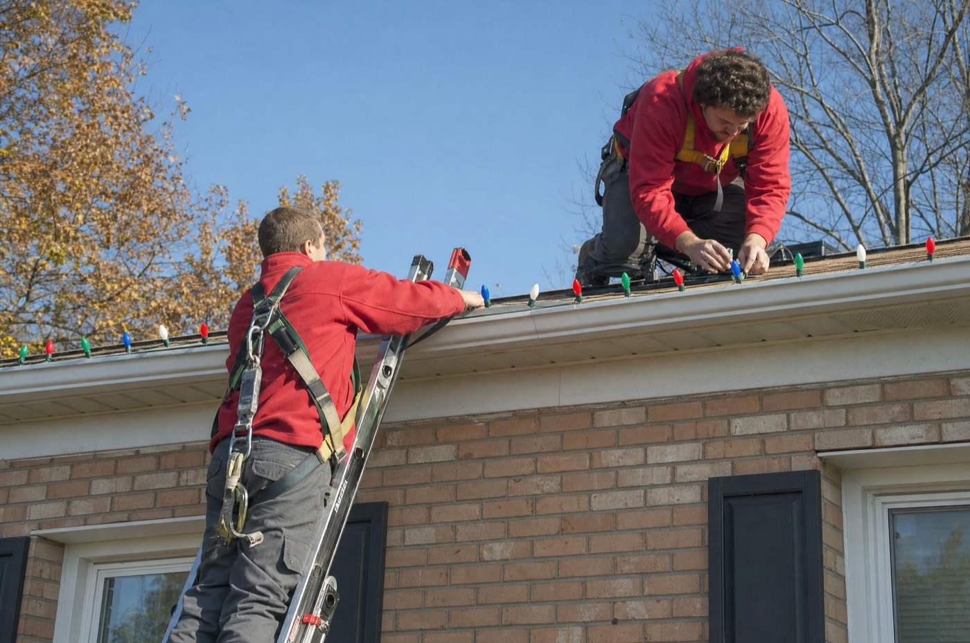 Two installers in harnesses clipping lights to a brick home's gutter on a sunny afternoon