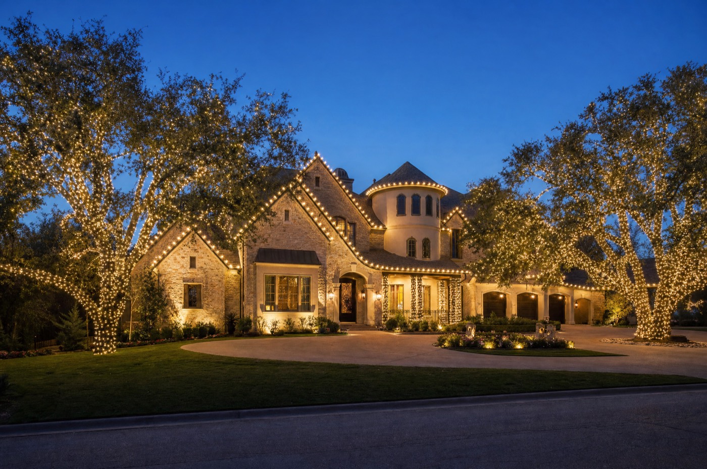 Large stone estate with every peak outlined in warm white lights and two wrapped live oaks