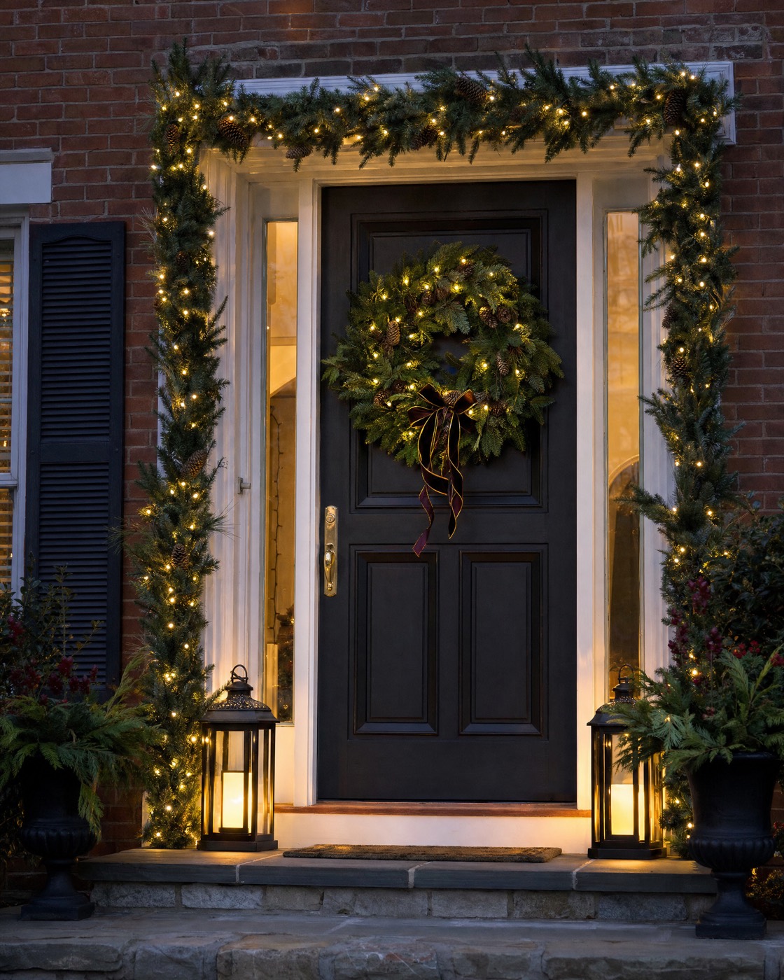 Front door framed in lit garland with a lit wreath and lanterns
